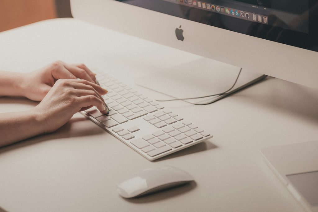 Fehér íróasztalon egy Mac billentyűzeten dolgozik két kéz, és kicsit bele lóg egy iMac